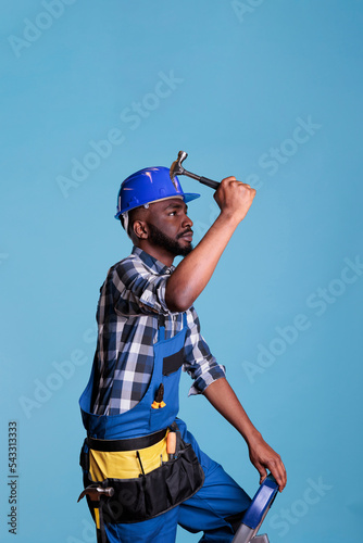 African american construction worker on ladder using hammer to drive nails into wall. Handyman wearing coveralls and protective helmet isolated on blue background in studio shot. © DC Studio