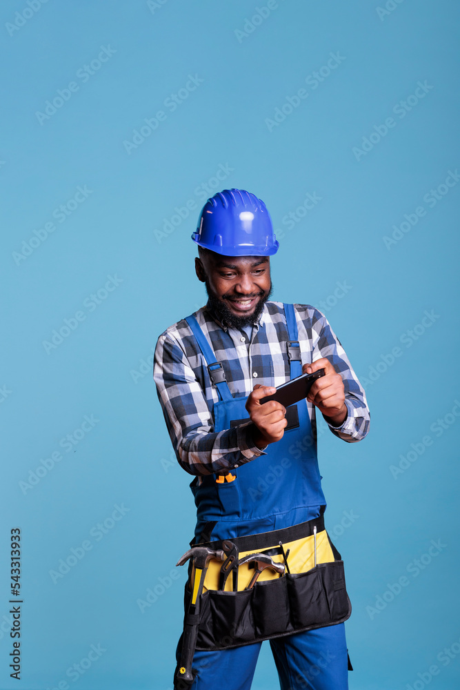 Contractor playing video games with mobile phone against light blue background in studio shot. Construction worker wearing coveralls and tool belt relaxing during break time from work. Contractor playing video games with mobile phone against light blue background in studio shot. Construction worker wearing coveralls and tool belt relaxing during break time from work.