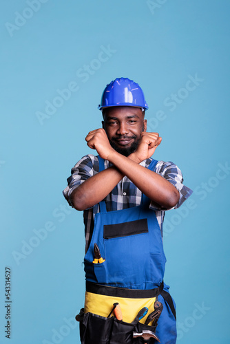 Disapproving african american builder making stop gesture with crossed arms while standing on blue background. Construction worker showing deny hands symbol while refusing to accept more work.