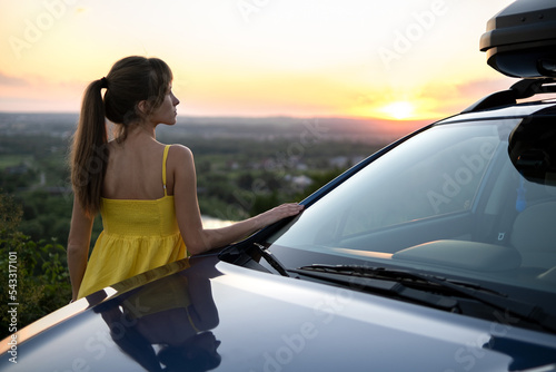 Happy young woman driver in yellow dress leaning on her car enjoying warm summer day. Travelling and vacation concept