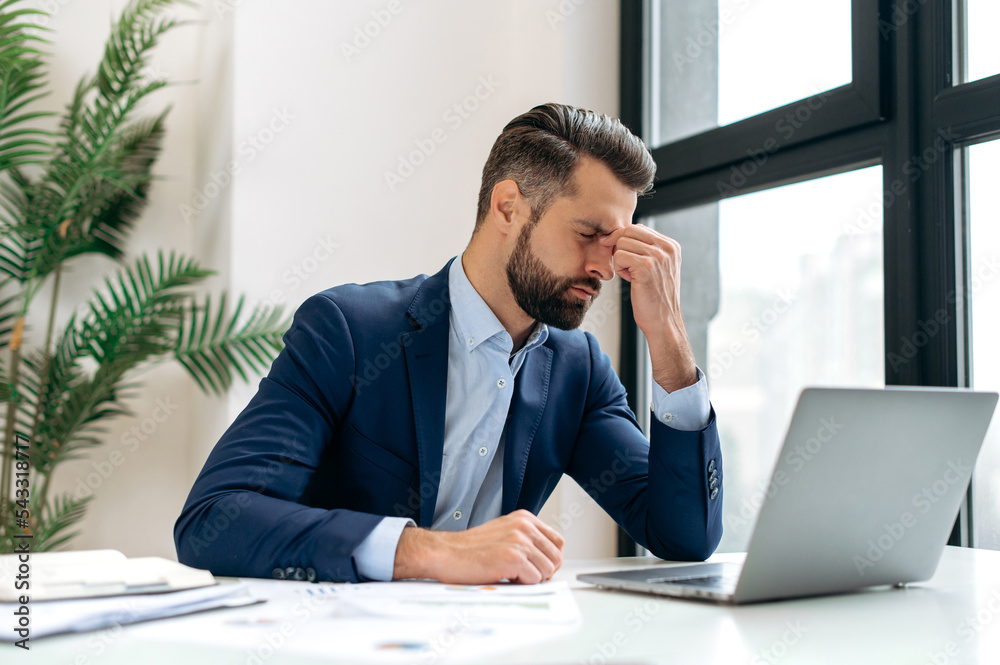 Frustrated overworked caucasian businessman, entrepreneur, product manager, programmer, sits at a desk in the office, massaging the bridge of his nose with his eyes closed, exhausted from long work Frustrated overworked caucasian businessman, entrepreneur, product manager, programmer, sits at a desk in the office, massaging the bridge of his nose with his eyes closed, exhausted from long work