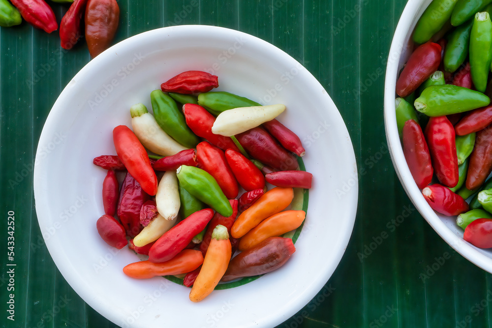 Small red, green, orange and creamy white chilli peppers in white dishes on a green banana leaf background. Philippines.