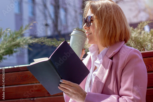 A young beautiful girl in sunglasses drinks coffee from an eco-friendly thermocup and holds a notebook while sitting on a bench outside in warm weather. photo