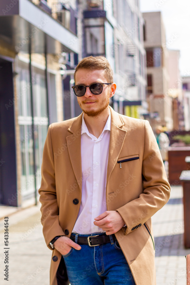 A young man dressed in a beige coat is walking down the street. A young bearded guy with a modern hairstyle and sunglasses on an urban background