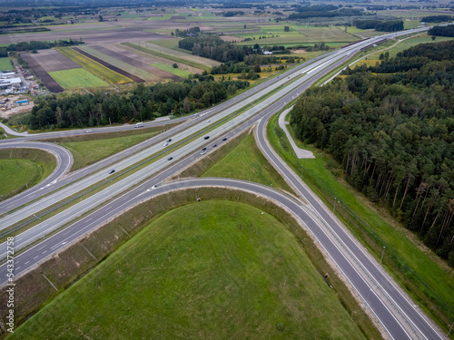 The picture presents intersection of roads visible from above with multiple cars and grass around.