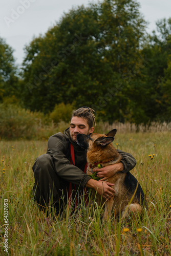 Concept of love and loyalty with animal. Male pet owner sits in grass in autumn and hugs dog. Young Caucasian man with dreadlocks and beard, German shepherd licks his face.
