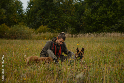 Male pet owner smiling, sitting in green grass and stroking Australian and German Shepherd with hands. Young Caucasian man with dreadlocks and beard sitting in autumn field with two purebred dogs. © Ekaterina