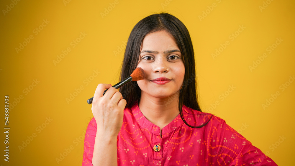 Beauty portrait of an attractive lovely young Indian woman isolated over yellow background, holding makeup blusher brush Beauty portrait of an attractive lovely young Indian woman isolated over yellow background, holding makeup blusher brush
