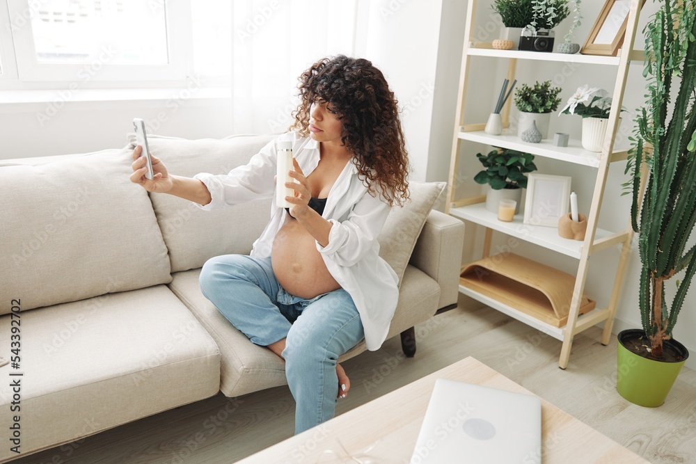 Pregnant woman smile blogger showing baby feeding bottle filming herself on phone, video chat sitting on sofa at home freelancer in last month of pregnancy Pregnant woman smile blogger showing baby feeding bottle filming herself on phone, video chat sitting on sofa at home freelancer in last month of pregnancy