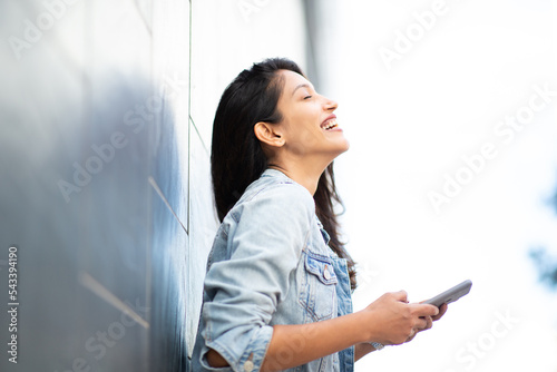 Side of laughing woman holding cellphone by wall