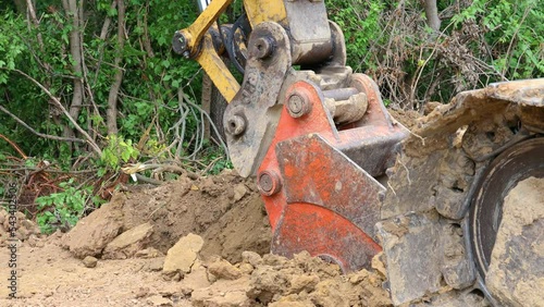 Close up of an with orange bucket of a hydraulic excavator digs an overflow drainage ditch at  pond construction site; a man with surveyor's tools checks the depth of the ditch photo