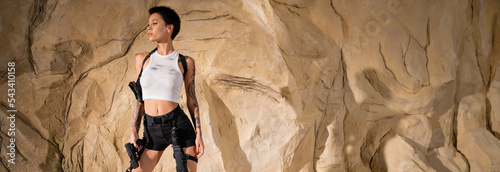 tattooed young archaeologist in sexy outfit holding gun and standing near rocks, banner. photo