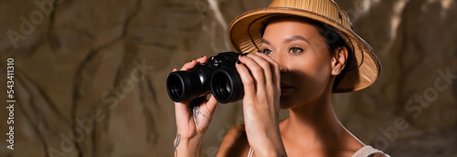 young and pretty archaeologist in safari hat holding binoculars and looking away in cave, banner. © LIGHTFIELD STUDIOS