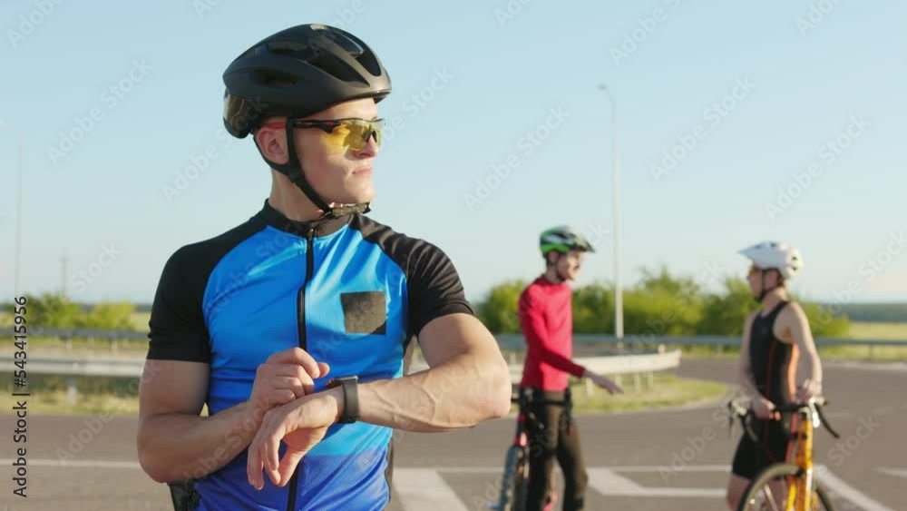 Male athlete in helmet and glasses using smartwatch while standing on road with colleagues on background. Professional cyclist checking heart rate and distance on modern gadget after workout. Male athlete in helmet and glasses using smartwatch while standing on road with colleagues on background. Professional cyclist checking heart rate and distance on modern gadget after workout.