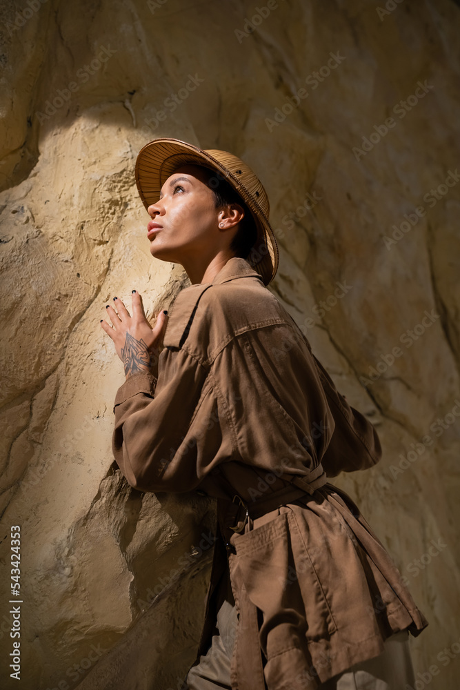 archaeologist in beige jacket and safari hat looking away near rock in cave.