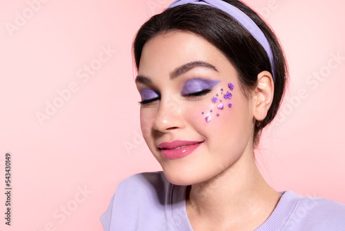 Stylish girl with shiny skin, lavender eyeshadow, spring flowers on cheek. Cool teen with glowing healthy skin, fresh complexion pose on pink studio background. Skincare cosmetics advertising