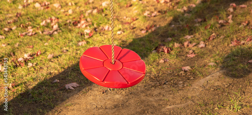 Red disc swing hanging from a rope. The shadow of a tree is behind it. To the right is the round shadow of the swing. Bare dirt is beneath the swing and red fall leaves are scattered on the ground.