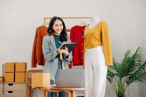 Fashion designer l young asian woman working using laptop, tablet and smiling while standing in workshop Responding on business e-mail..