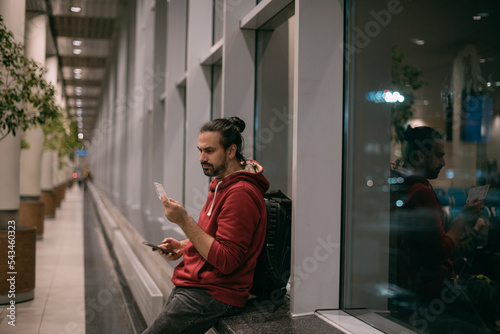 Obraz na plátně A young man has checked in online for a plane, is waiting for a plane with a boarding pass in his hands, with hand luggage at the airport at the gate