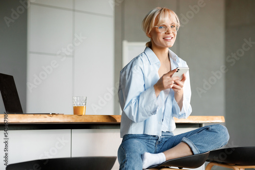 Beautiful young blond girl in stylish glasses is working on her phone. Smiling and looking at the camera.