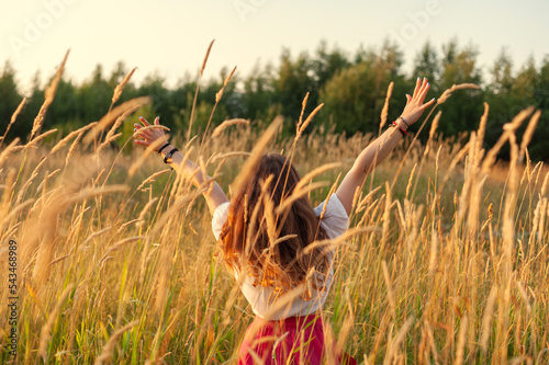 young girl who looks like hippie standing with her back to the camera on field at sunset raises her hands up