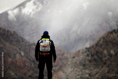 A man with backpack stands on a mountainside and looks into the distance at a mountain peak. A lone hiker travels high in the mountains on a cloudy day. Independent hike in Pamir mountains. 