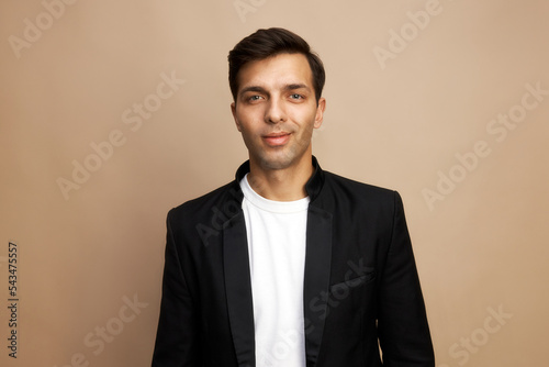 Portrait of smiling successful young handsome businessman participating in tender process, isolated on brown studio background in white t-shirt and black jacket, feeling himself confident