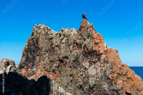 Horizontal photo of a man sitting on high cliffs of red granite rock in the peninsula of Kullaberg, Skåne, Sweden. Man climbing cliffs next to the baltic ocean, looking out towards the sea on the top