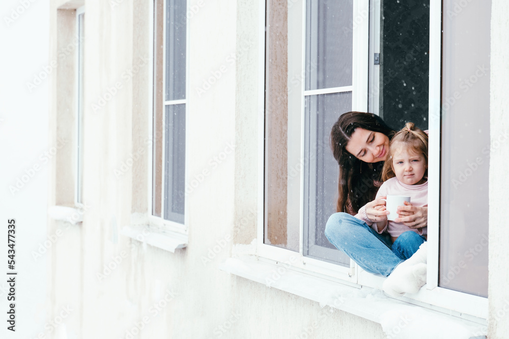 Cute portrait of mother and daughter sitting on a window sill at home and enjoying winter snowy weather. Cute portrait of mother and daughter sitting on a window sill at home and enjoying winter snowy weather.