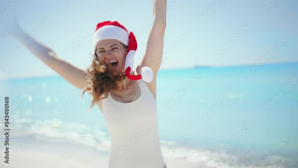 smiling modern 40 years old woman in white swimsuit with long wavy hair and striped christmas hat jumping at the beach.
