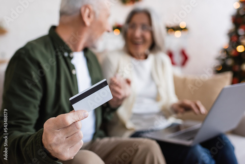 mature man holding credit card near wife with laptop while having online shopping on blurred background