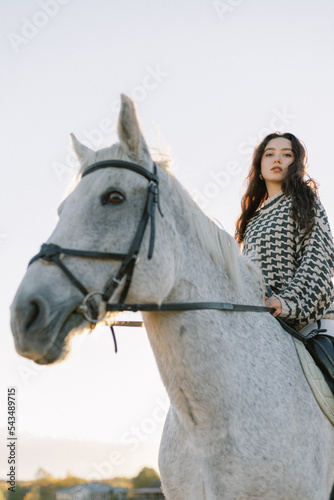 A young brunette and a white horse on a ranch. Rider on a horse at sunset. Pet, domestic-bred farm animals. Feeling free. 