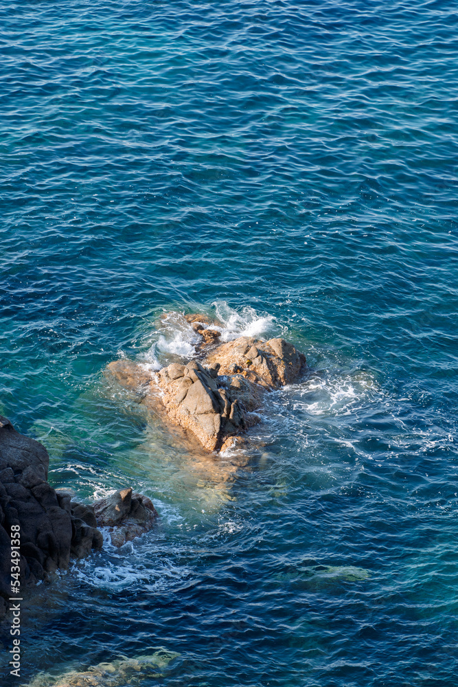 Obraz premium Wild rocky beach coastline and the sea, wave splashes on rock in sea shore, calm sea. Breaking waves to the rocks on shore creating white sea foam and small bubbles, top view