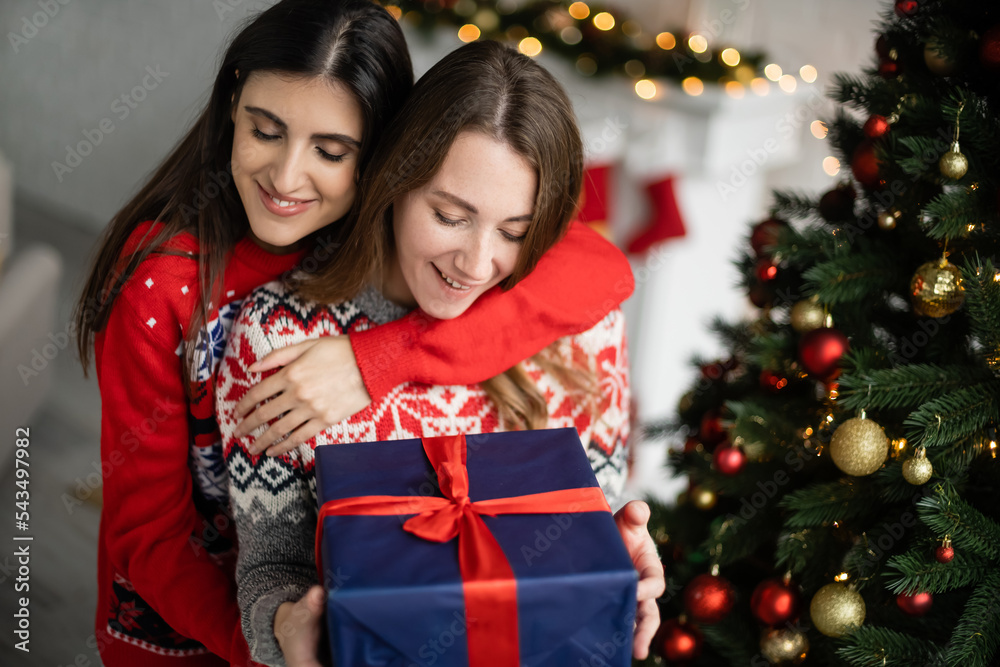 Positive woman hugging girlfriend with present near christmas tree at home Positive woman hugging girlfriend with present near christmas tree at home