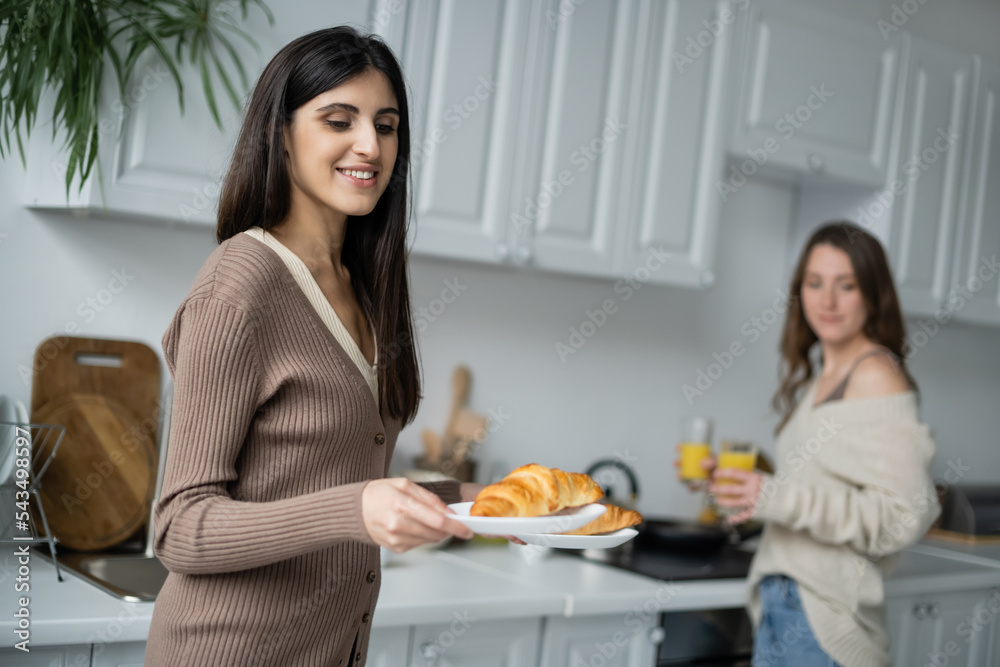 Lesbian woman holding croissants near blurred girlfriend in kitchen