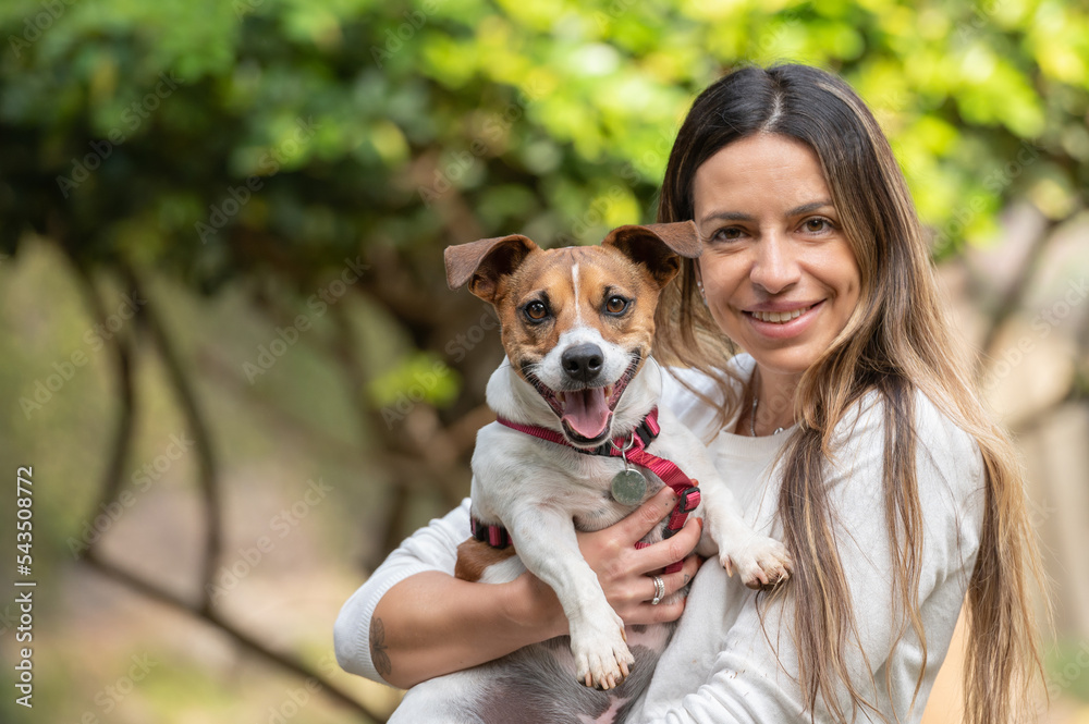 Woman holding Jack Russell dog posing for the camera at the park during the day Woman holding Jack Russell dog posing for the camera at the park during the day