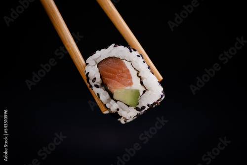 salmon cheese and avocado uramaki roll held by chopsticks on a black background