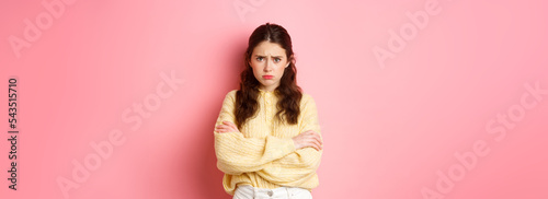 Cute silly girl feeling offended and sad, trying to comfort herself with self hug, frowning and looking at camera insulted, about to cry, standing against pink background