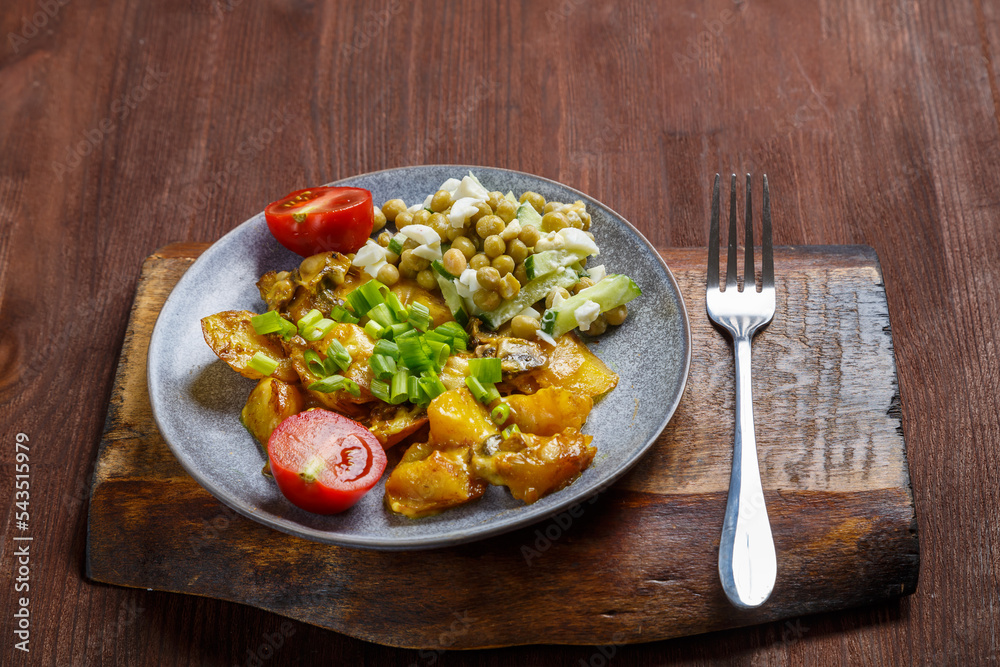 Potatoes baked with cheese and mushrooms garnished with cherry tomatoes sprinkled with green onions in a plate on a wooden board next to a fork.