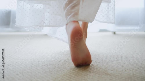 Back view of woman in white dress, nightie walking barefoot at home. Female go on tiptoe to the window along the carpet in the morning