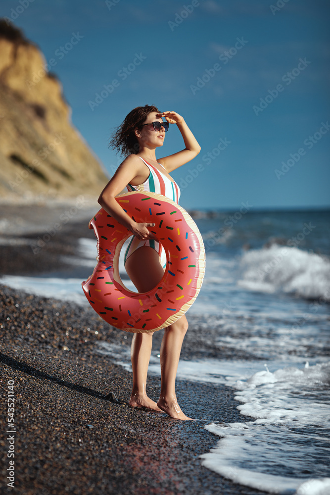 Tanned young woman in a swimsuit and sunglasses standing on a beach with an inflatable ring and looking at the see.