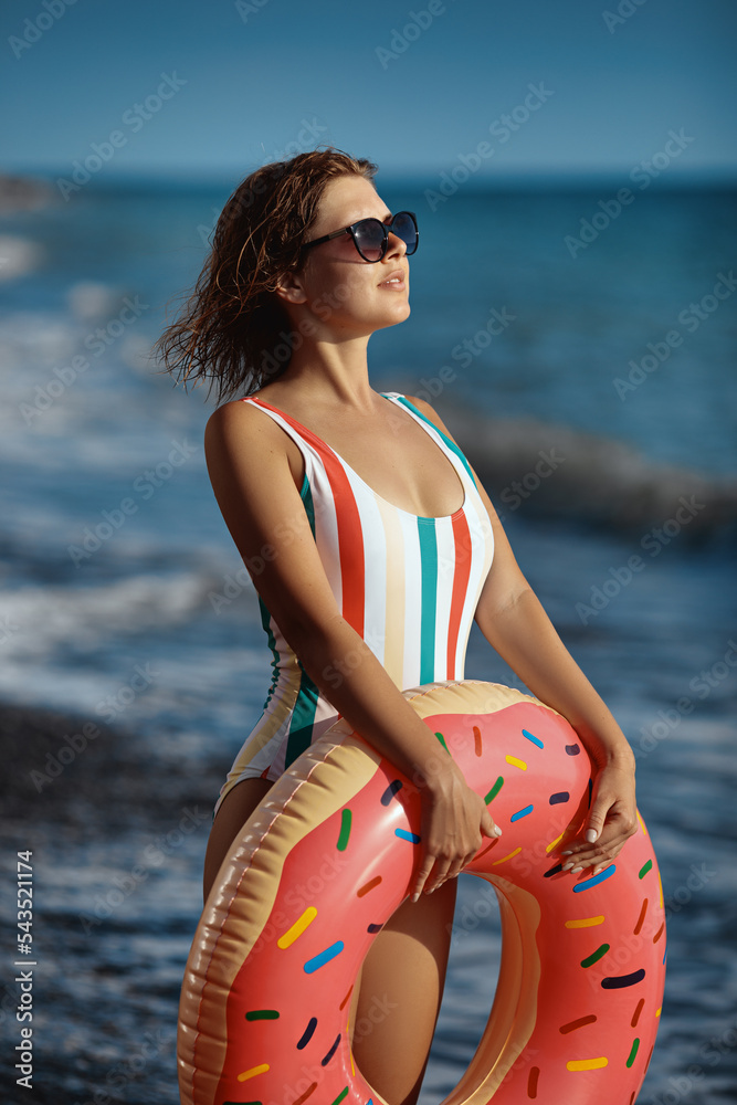 Smiling young woman in a swimsuit and sunglasses standing on a beach with an inflatable donut ring and enjoying sunbathing.