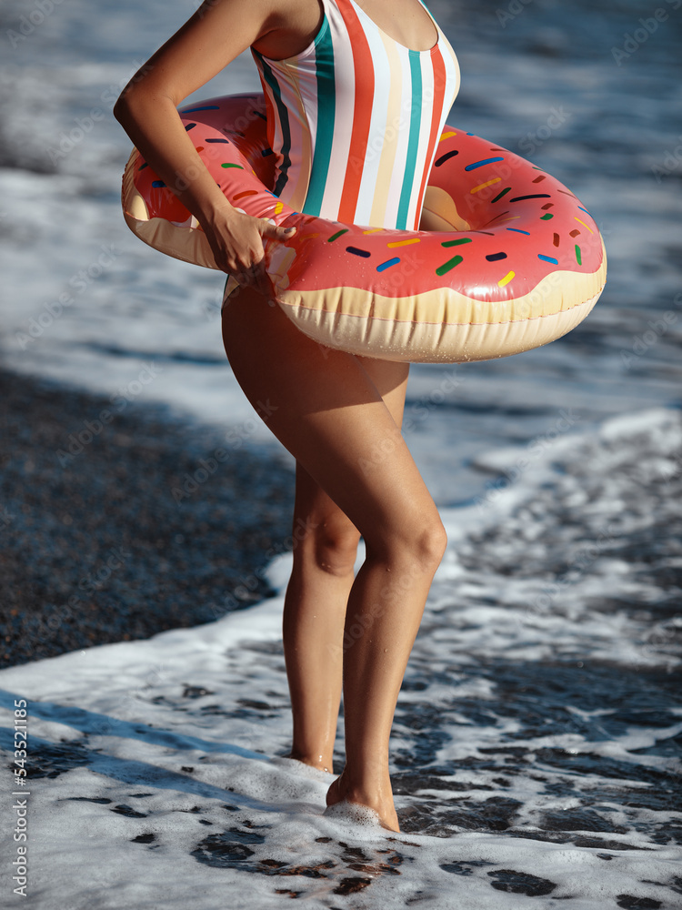 Young woman in a swimsuit standing on a beach with an inflatable donut ring and enjoying sunbathing.