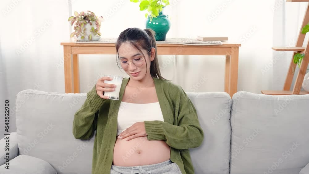 4K, A pregnant Asian mother with her maternity tummy sits on the sofa in the living room of her home drinking milk and stroking her tummy, showing warmth to her upcoming baby. 4K, A pregnant Asian mother with her maternity tummy sits on the sofa in the living room of her home drinking milk and stroking her tummy, showing warmth to her upcoming baby.