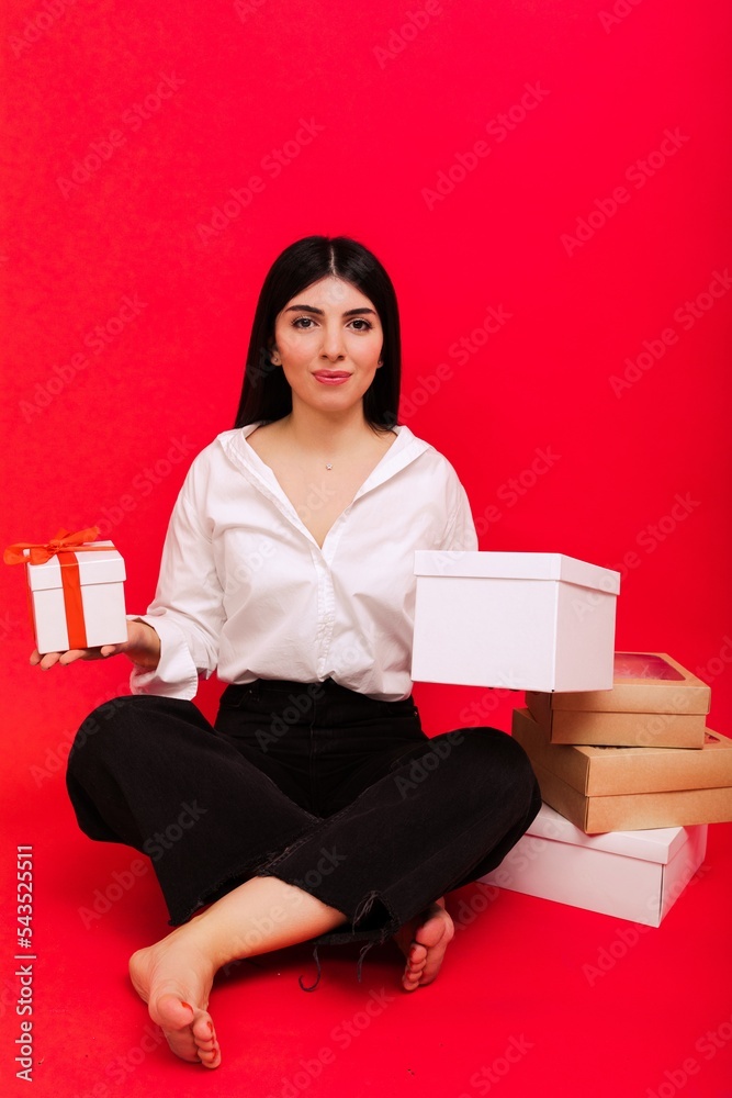 Christmas and New Year. Portrait of a young woman in a white shirt and jeans holding boxes with gifts and sitting on a red background in the studio Christmas and New Year. Portrait of a young woman in a white shirt and jeans holding boxes with gifts and sitting on a red background in the studio
