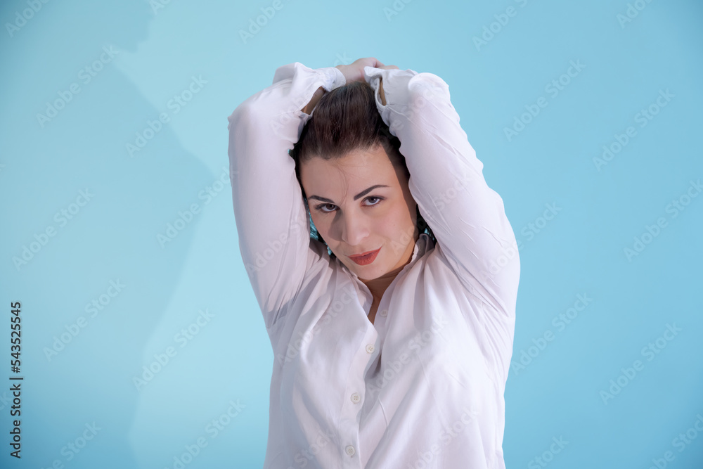 Close-up portrait of beautiful thoughtful caucasian brunette woman in white shirt with raised arms. Blue background. Selective focus. People theme. Close-up portrait of beautiful thoughtful caucasian brunette woman in white shirt with raised arms. Blue background. Selective focus. People theme.