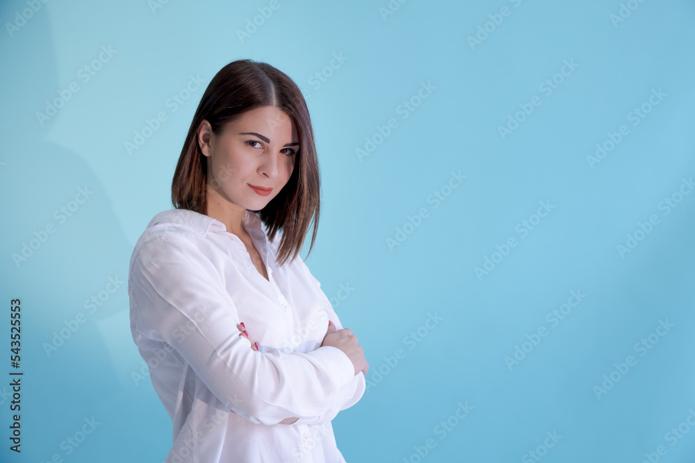 Close-up portrait of beautiful sensual caucasian woman with brown hair wearing white shirt and standing with crossed arms. Blue background. Selective focus. Copy space for your text. People theme. Close-up portrait of beautiful sensual caucasian woman with brown hair wearing white shirt and standing with crossed arms. Blue background. Selective focus. Copy space for your text. People theme.