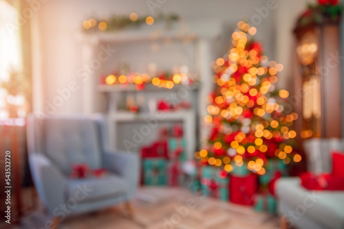Blurred view of modern Christmas room interior with light bokeh background. Decorated glowing tree  armchair  fireplace with candles and gifts
