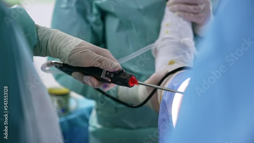 Gloved hand of a surgeon holding a device and using it at operation. Close up. Assisting medic holding a patient’s hand at backdrop. photo