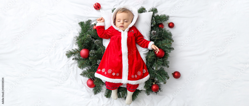 Cute little baby girl in Santa Claus costume and with Christmas decor lying on bed, top view